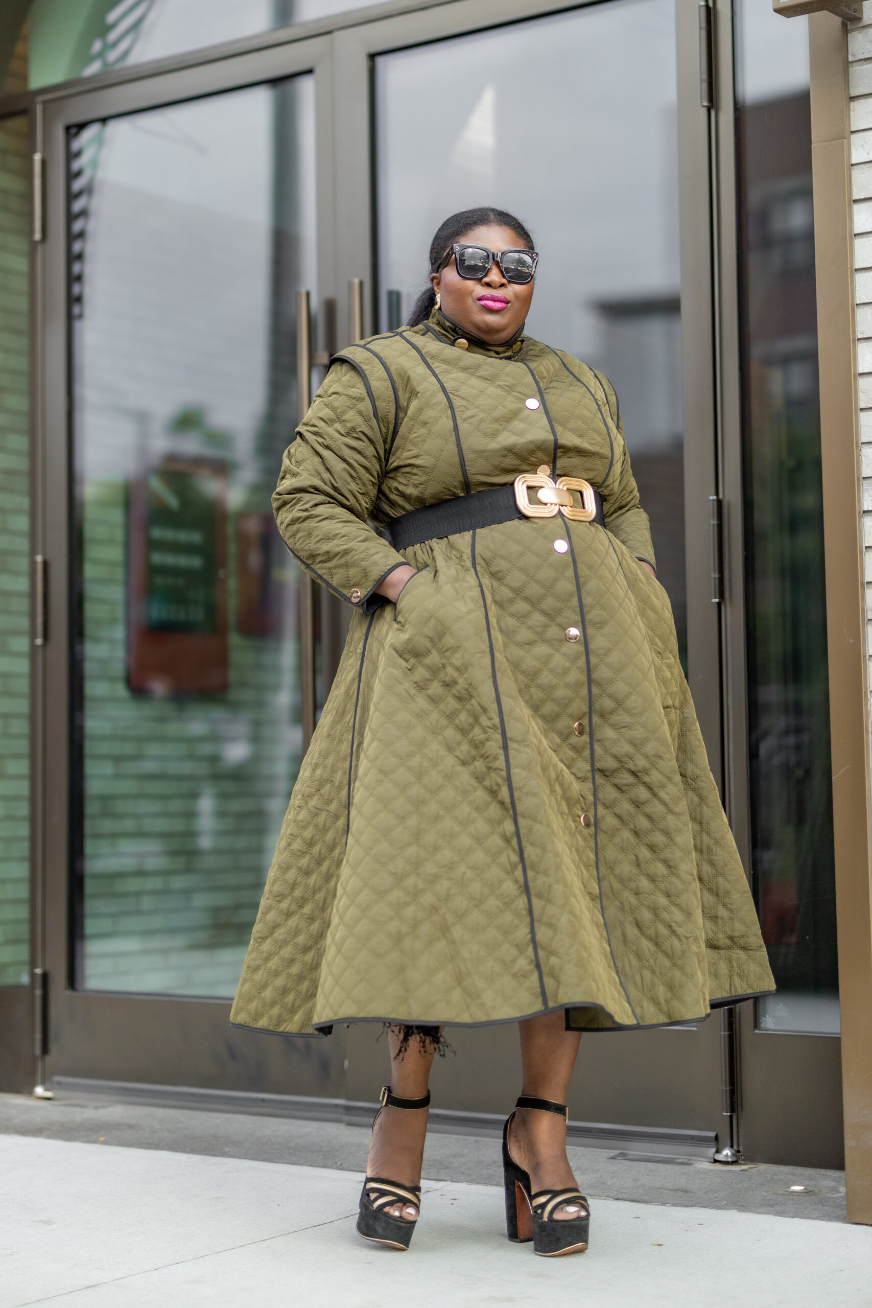 A Black woman wearing a quilted green coat dress with a black belt around the waist and black platform sandals. She's wearing sunglasses
