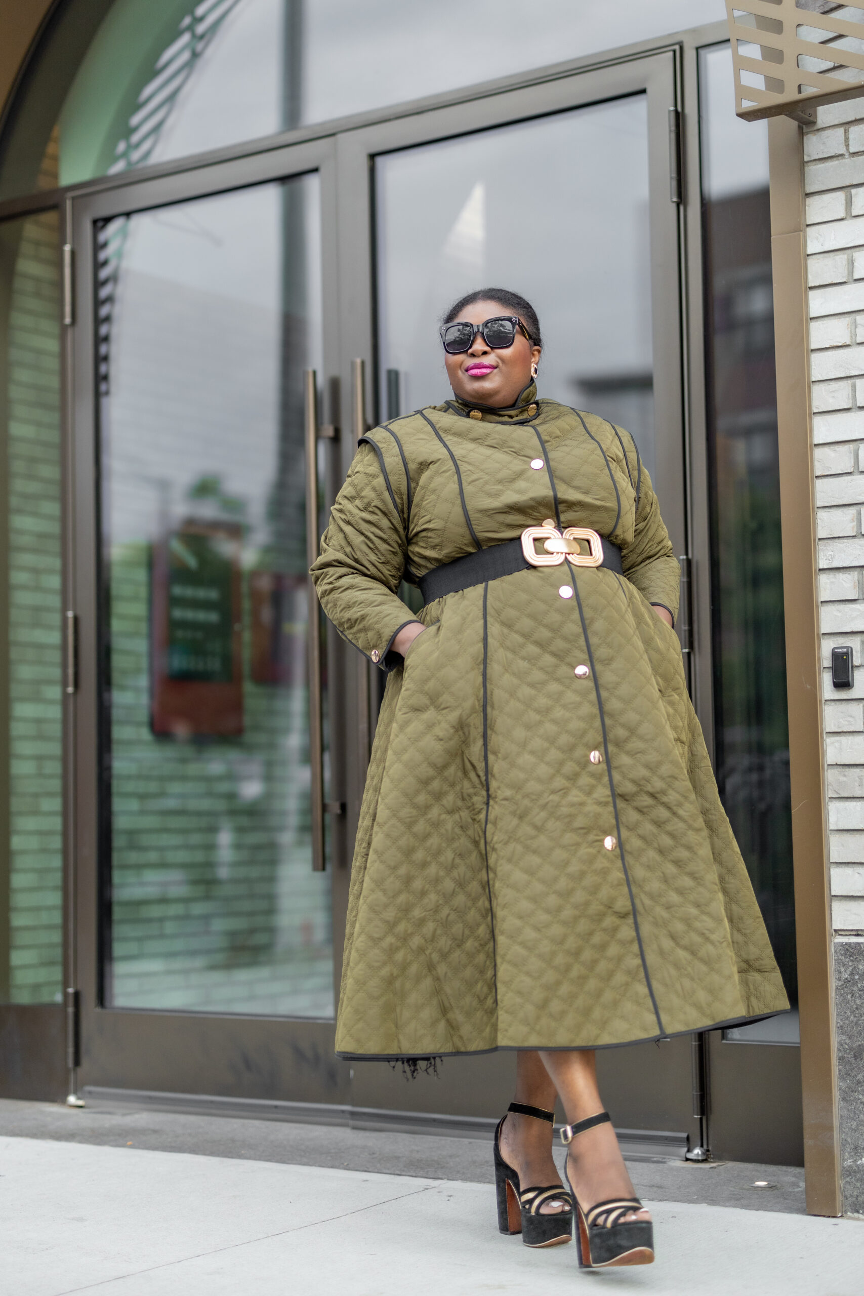 A Black woman wearing a quilted green coat dress with a black belt around the waist and black platform sandals. She's wearing sunglasses