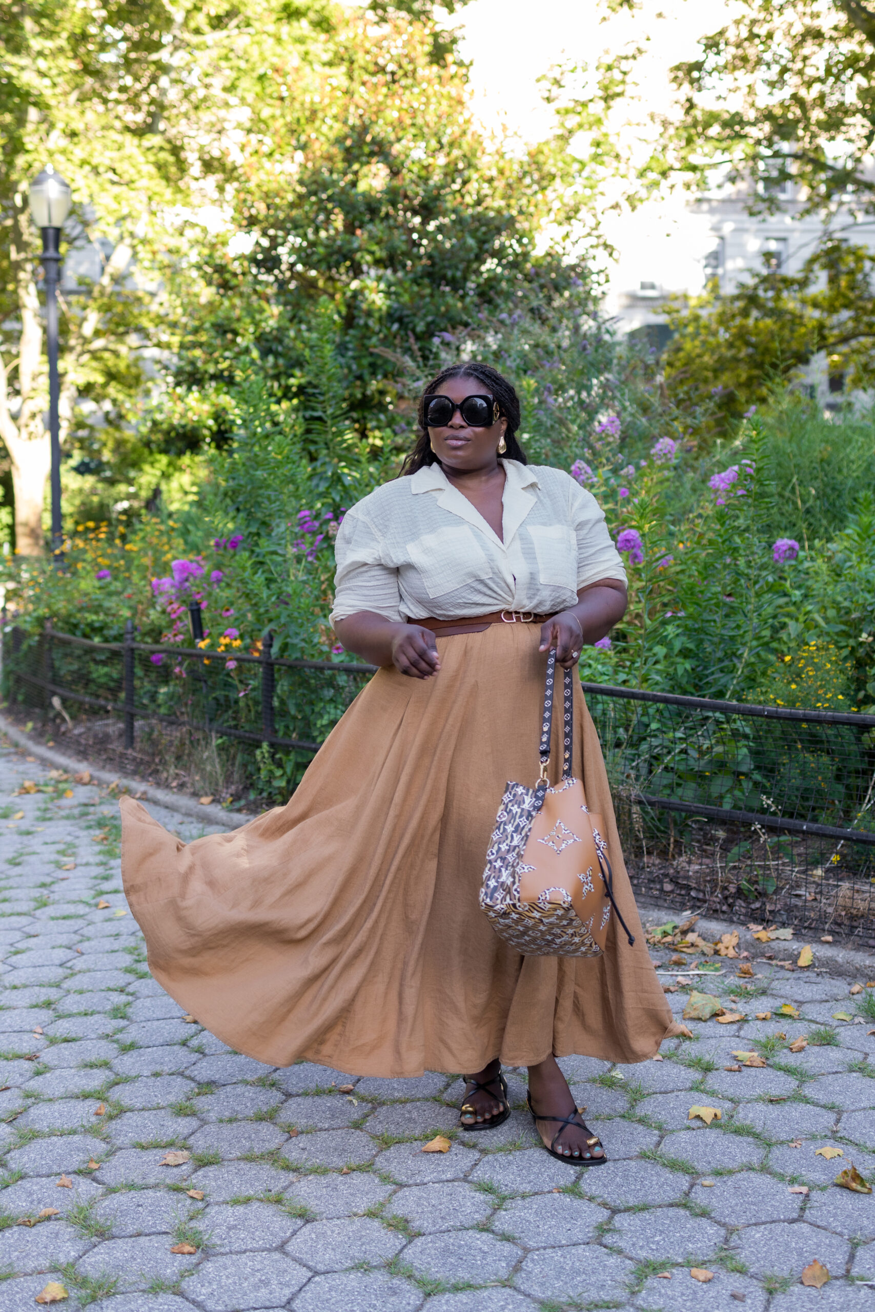 A Black woman wearing a cream button down shirt and a billowy long brown skirt, black sandals, and sunglasses in a city garden on a bright summer afternoon